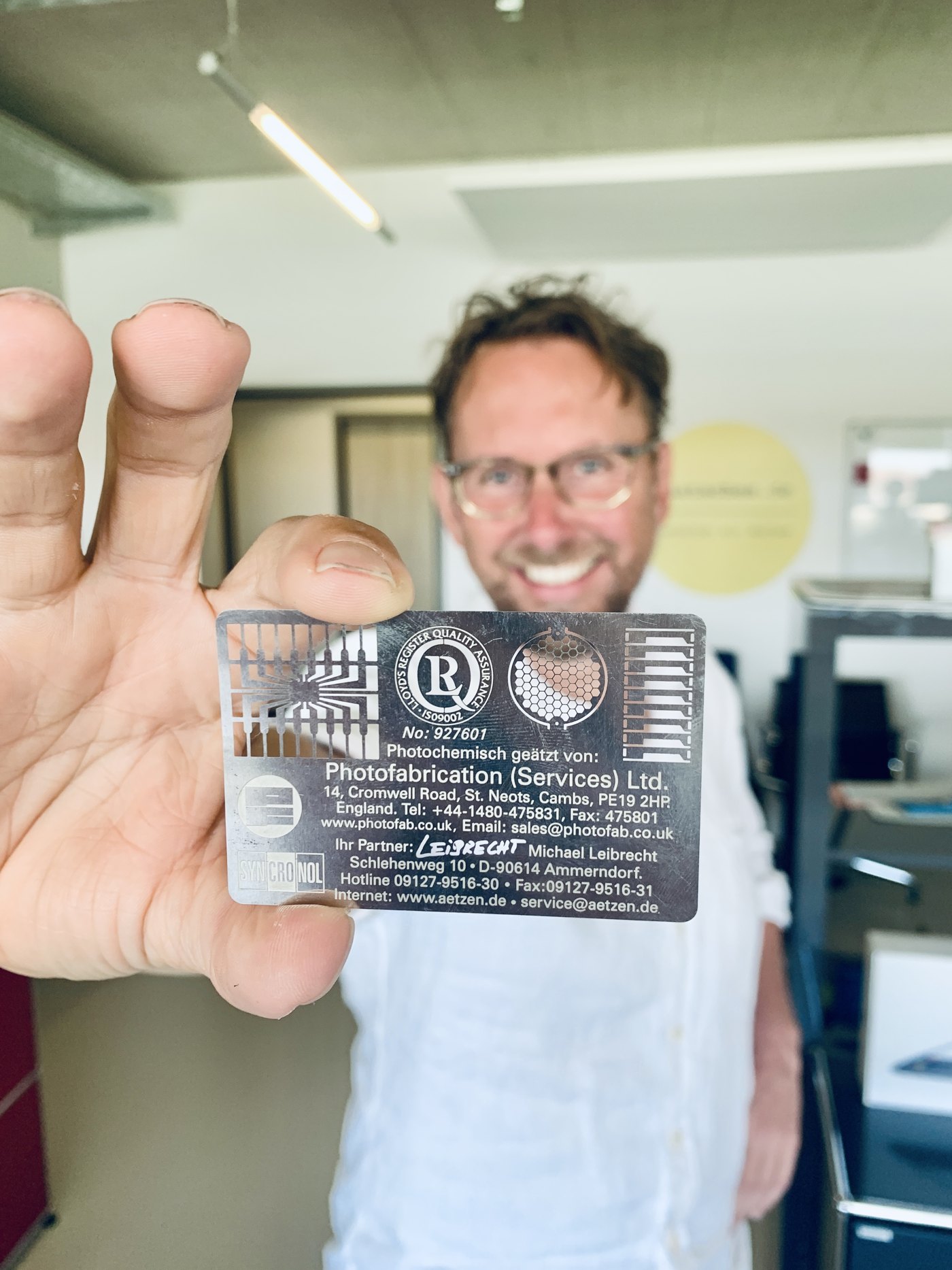 Michael Leibrecht hält eine photochemisch geätzte Edelstahl-Visitenkarte in die Kamera. Auf der Karte sind Ätz-Demonstrationsstrukturen zu sehen sowie die Aufschrift „Photochemisch geätzt von: Photofabrication (Services) Ltd., England
